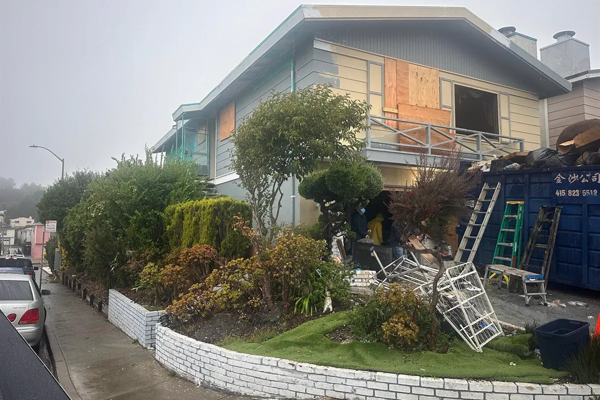 The same corner home before the rebuild, with boarded windows, stripped siding and a dumpster in the driveway
